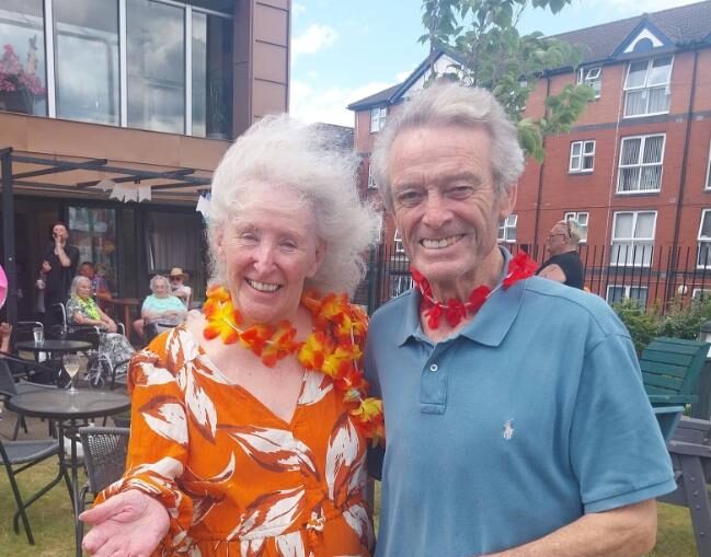 two older adults stood in the garden with flower necklaces