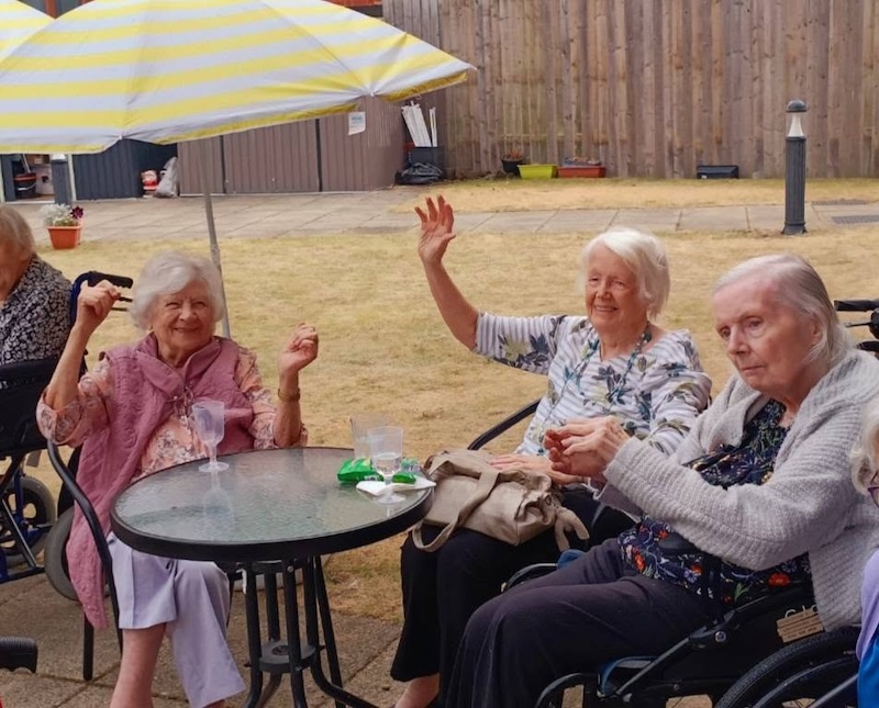 2 female residents sat together in the garden smiling with their hands up